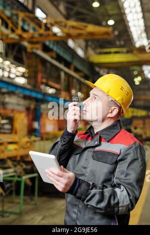 Portrait of mature manual worker in helmet and workwear looking at camera while using tablet pc in the factory Stock Photo
