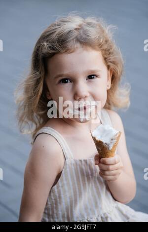 Toddler child eating cone ice cream outside near cafe. Cute blonde Caucasian girl licking frozen summer food on the street. Dirty mouth, happy kid. Stock Photo