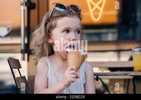 Toddler child eating cone ice cream outside near cafe. Cute blonde Caucasian girl licking frozen summer food on the street. Dirty mouth, happy kid. Stock Photo