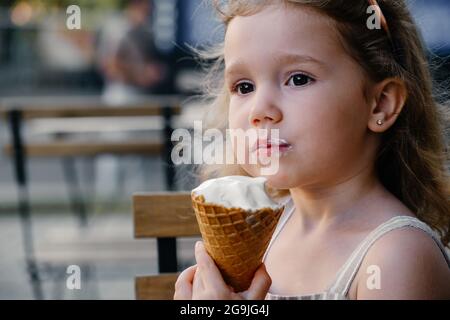 Toddler child eating cone ice cream outside near cafe. Cute blonde Caucasian girl licking frozen summer food on the street. Dirty mouth, happy kid. Stock Photo