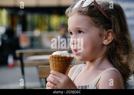 Toddler child eating cone ice cream outside near cafe. Cute blonde Caucasian girl licking frozen summer food on the street. Dirty mouth, happy kid. Stock Photo