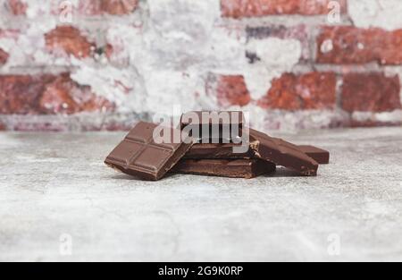 On a gray surface in front of a brick wall are several pieces of a dark chocolate bar stacked up. Stock Photo