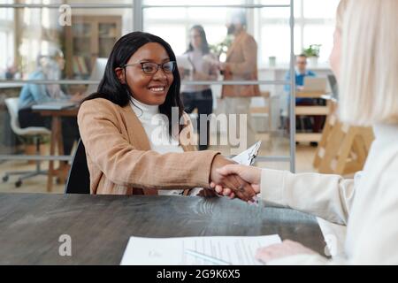 Young cheerful African female specialist shaking hand of new employer after interview Stock Photo