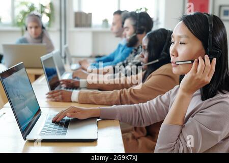 Young Asian operator consulting clients online against her co-workers in office Stock Photo