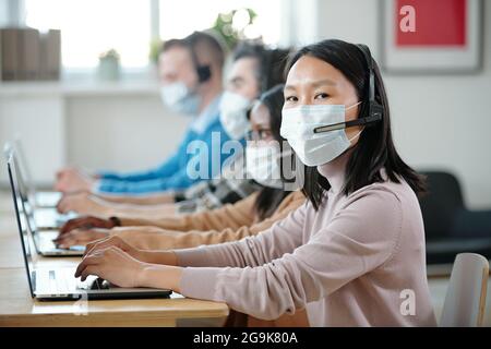 Young Asian operator in protective mask looking at camera against co-workers Stock Photo