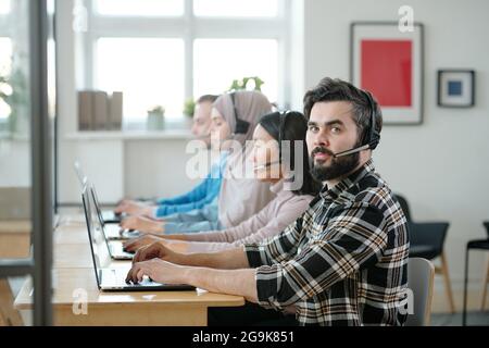 Bearded businessman in headset looking at you during work with online clients among co-workers Stock Photo
