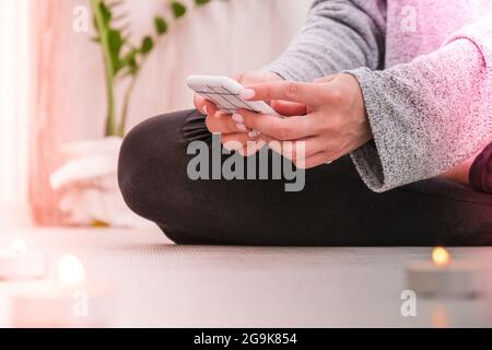 Young millennial blonde woman doing yoga exercise watching tutorial lesson on mobile phone stretching fitness at balcony home. Mindfulness meditation. The concept of online training. Video lessons. Self-isolation is beneficial. Exercise for Lose weight, increase flexibility And tighten the shape. Young slim athletic girl practices yoga at home in online lessons. Stock Photo