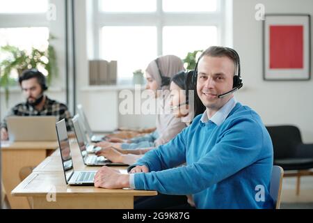Young successful operator with headset looking at you with smile against his co-workers Stock Photo