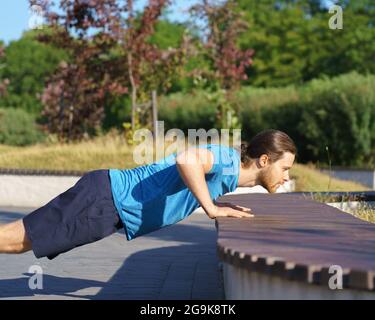 Sportive guy training arms while doing push ups on bench in park on summer day Stock Photo