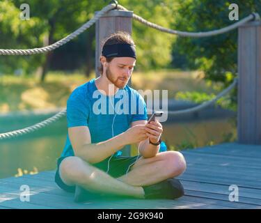 Calm young sportive man in earphones using modern smartphone while resting after exercising in park Stock Photo