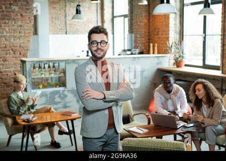 Young smiling cross-armed businessman standing in front of working colleagues Stock Photo