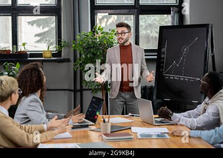 Confident business coach explaining graph by blackboard to young intercultural managers Stock Photo