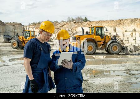 Two builders in workwear looking at sketch of building on tablet screen Stock Photo