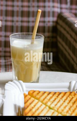 A glass of coffee drink frappe, cappuccino, latte with an eco-friendly paper tube and two grilled sandwiches on a beautiful white plate against a chec Stock Photo