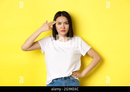 Woman showing kill me please gesture, shooting herself in head with finger gun from boredom, standing upset over yellow background. Stock Photo