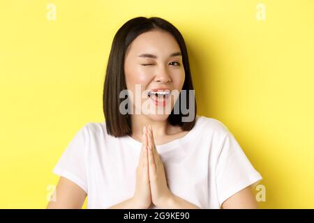 Close of cheeky young asian woman holding hands in namaste, thank you gesture, winking at camera coquettish, feeling lucky, standing over yellow backg Stock Photo