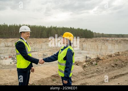 Two colleagues in hardhats and workwear shaking hands against quarry Stock Photo