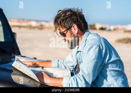 Man look a paper road trip map standing outside the car with blue sky and ground desert in background - concept of travel and adventure lifestyle peop Stock Photo