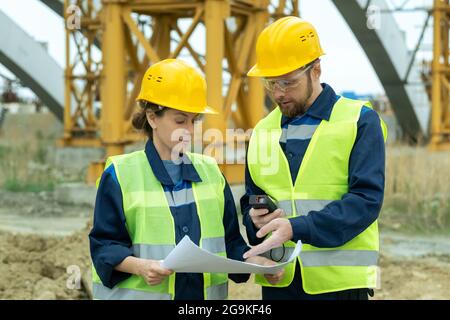 Young man pointing at blueprint and discussing with his colleague the future project on construction site Stock Photo