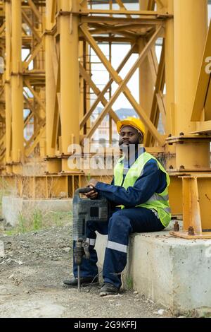 Portrait of African construction worker in workwear looking at camera while sitting outdoors with work tool Stock Photo