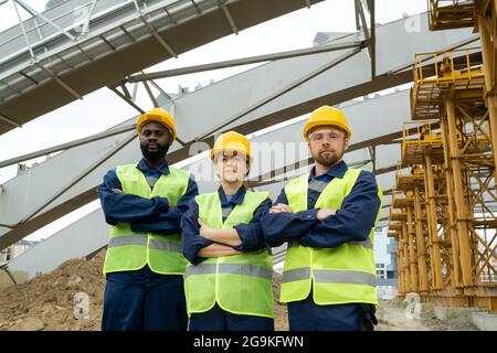 Portrait of group of engineers in reflective clothing and work helmets standing with arms crossed and looking at camera while standing outdoors Stock Photo