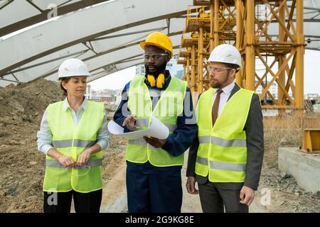 Group of engineers examining blueprint and discussing new construction project in team while walking through construction site Stock Photo