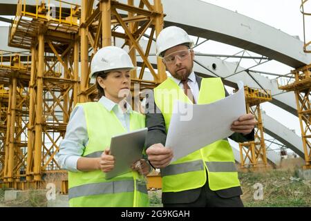 Young architect in work helmet examining blueprint and discussing it with his colleague while she using tablet pc outdoors Stock Photo