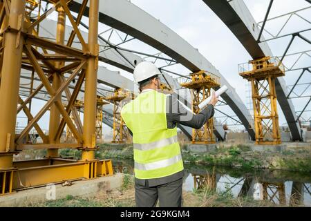 Rear view of engineer with blueprint looking at construction building and talking on mobile phone outdoors Stock Photo