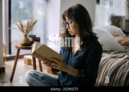 Young focused asian woman wearing eyeglasses reading book while sitting in lotus pose on floor in cozy living room Stock Photo