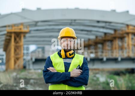 Portrait of foreman in headphones and in work helmet standing with arms crossed on construction site and looking at camera Stock Photo