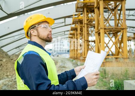 Engineer in workwear examining blueprint and looking at construction of the building outdoors Stock Photo