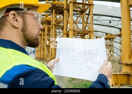 Architect in work helmet examining the blueprint of future project while working on construction site Stock Photo