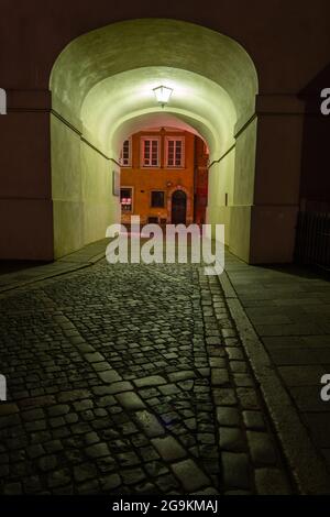 Cobblestone empty street at night with arched passage under building, illuminated with single lamp in Old Town of Warsaw, Poland. Stock Photo