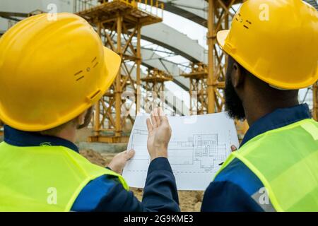 Rear view of two workers in work helmets comparing the blueprint with the building under construction while standing outdoors Stock Photo