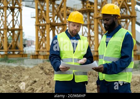 Two engineers in reflective clothing examining blueprint and discussing the project together while working on construction site Stock Photo