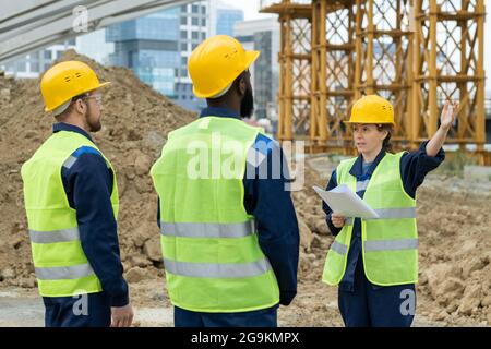 Foreman with blueprint pointing at construction and talking to workers during their work outdoors Stock Photo