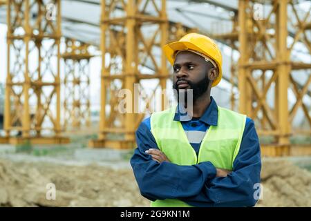 African construction worker in work helmet standing with arms crossed on the construction site and looking away Stock Photo