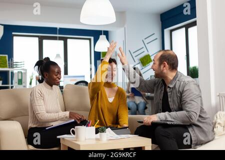 Diverse team of successful young entrepreneurs sharing high-five celebrating success sitting on couch in start up office room. Multiethnic business team with laptop and papers excited about project Stock Photo