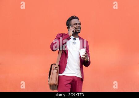 stylish african businessman dressed in suit smiles with a coffee on his hand while talks by phone in front of red background, copy space for text, con Stock Photo