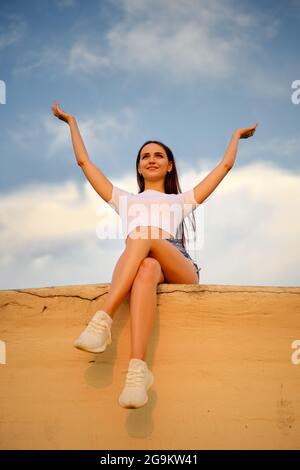 Smiling woman sit on a pier and raises her hands up Stock Photo