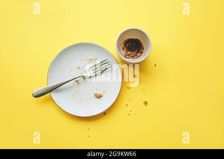 Empty plate with fork and crumbs after eating and cup with coffee grounds on yellow background. Concept of the end of the party or celebration. Stock Photo