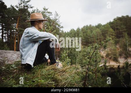 Side view low angle of young male traveler sitting on stony edge and admiring scenery of lake surrounded by coniferous forest Stock Photo