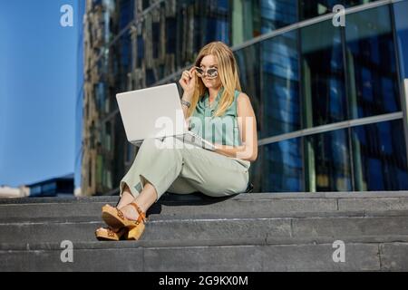 Manager with laptop is sitting on stairs near business center. Stock Photo