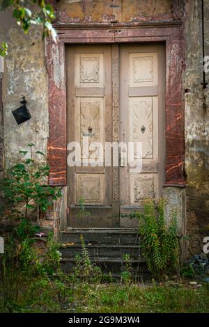 old, richly decorated but dilapidated wooden entrance door on an abandoned, enchanted-looking house with overgrown stairs and damage to the facade Stock Photo
