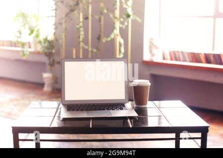 Photo of white blank screen of laptop with keyboard case lying on wooden little table with disposable cup of coffee from cafeteria next to computer ov Stock Photo