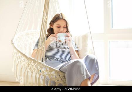 Beautiful young brunette woman drinking tea or coffee holding cup with both hands while relaxing in knitted macrame style rocking chair at home on sun Stock Photo