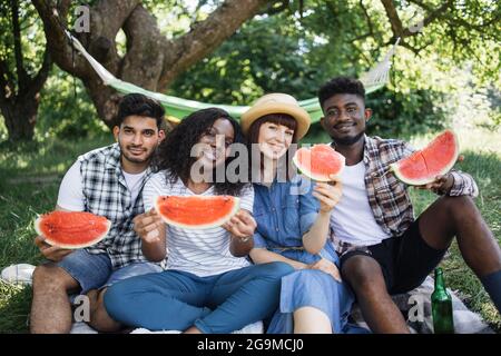 Four cheerful multiracial friends sitting at green garden with slices of sweet watermelon in hands. Young people having picnic on fresh air with tasty food. Stock Photo