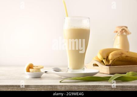 Milkshake with banana in glass cup on table with tray with fruit and white isolated background. Front view. Stock Photo