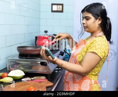 A pretty Indian young woman wearing apron watching cooking video in smart phone in domestic kitchen on gas stove Stock Photo