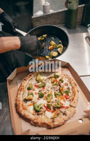 Close-up. Chef putting fried vegetables over hot tasty pizza in a cardboard box. Looks tasty, delicious Stock Photo